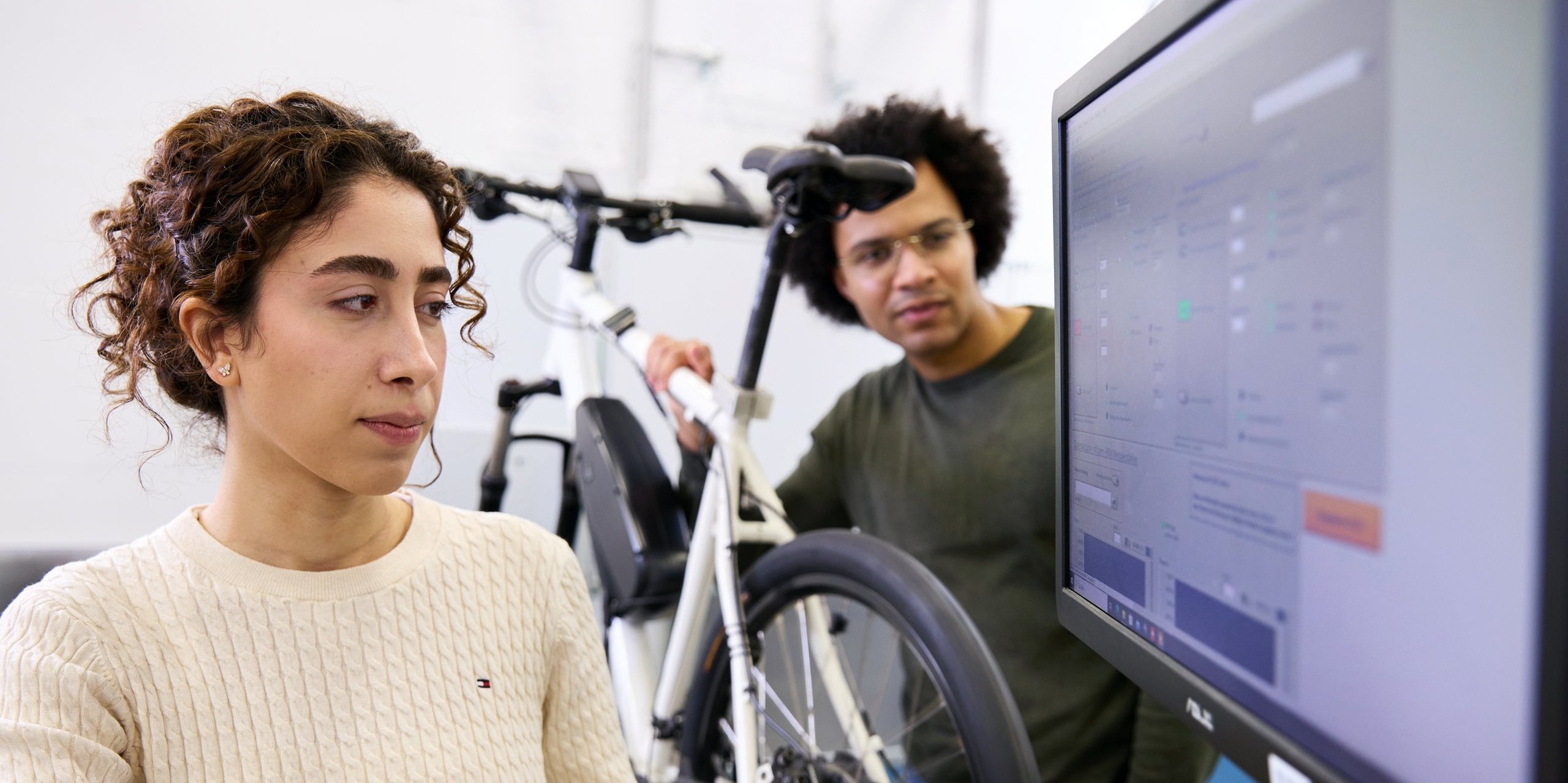 Person im Vordergrund auf BIldschirm schauend, Person im Hintergrund an E-Bike