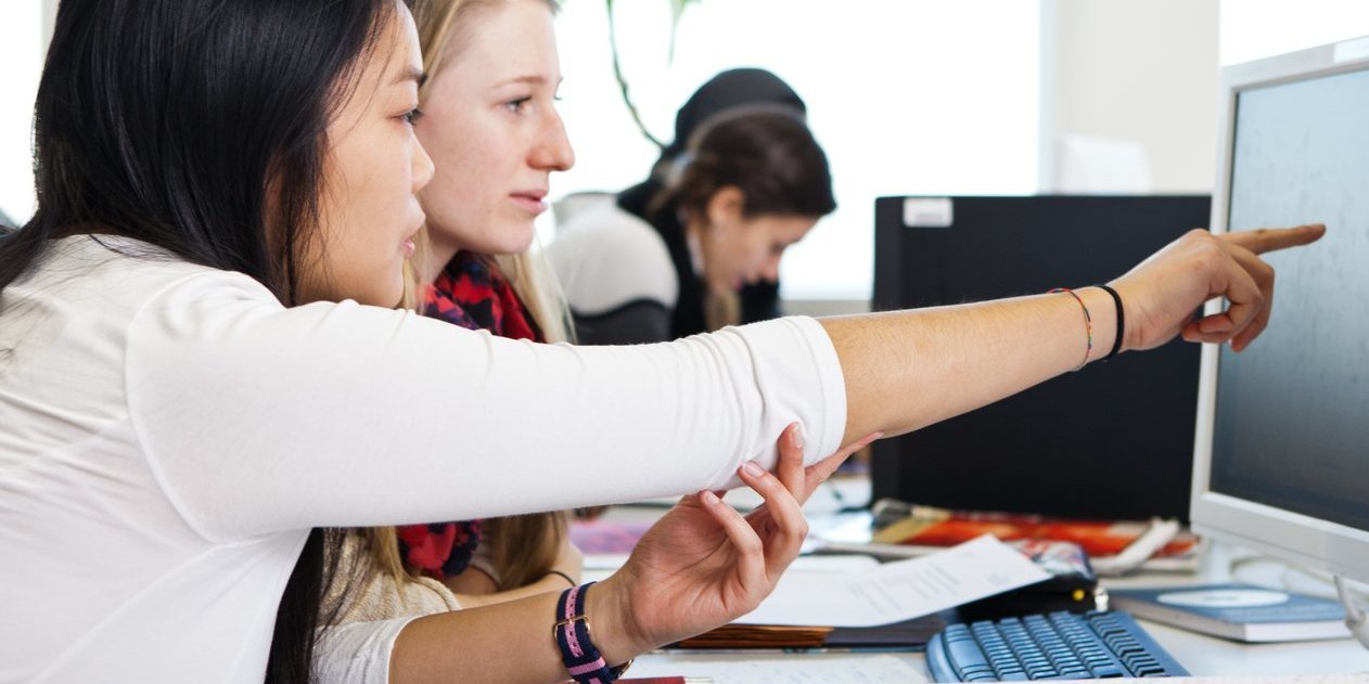 Eine Studentin erklärt einer anderen Studentin etwas und zeigt dabei auf den PC-Bildschirm.