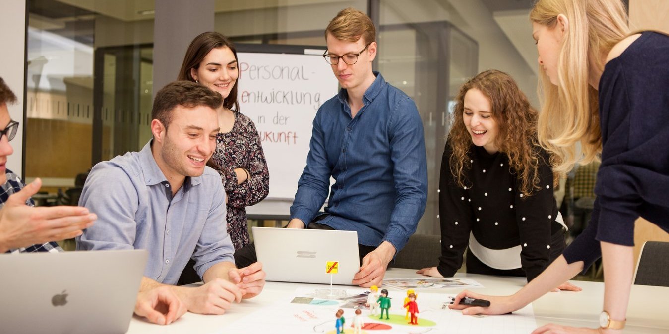 Auf dem Bild ist eine Gruppenarbeit zu sehen. Mehrere Studierende stehen um einen Tisch herum und arbeiten an einem Projekt.