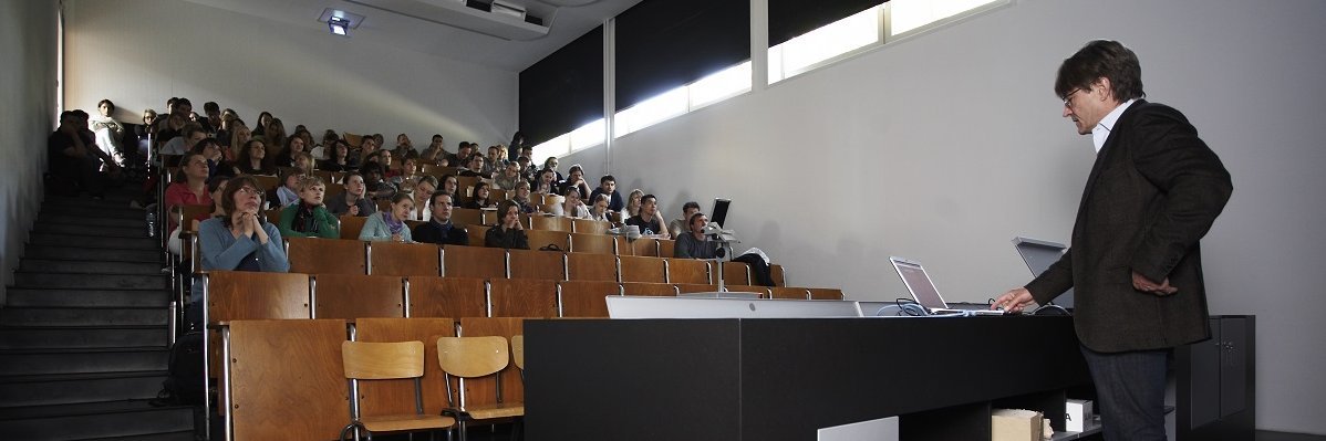 Vorlesung in einem leicht abgedunkelten, in Schwarz- und Holztönen gehaltenen modernen Stufenhörsaal.