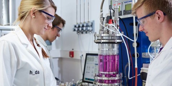 Mehrere Personen im Labor in weißen Laborkitteln und Schutzbrillen, die an einem chemischen Reaktor mit pinker Flüssigkeit und Mess-/Steuertechnik arbeiten