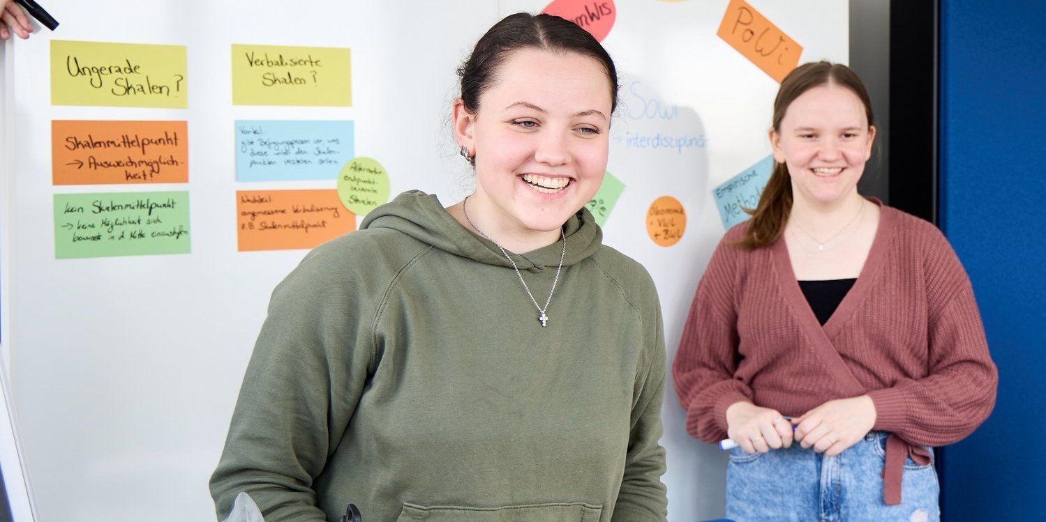 Zwei lächelnde Studentinnen stehen vor einer Wand mit bunten Notizzetteln