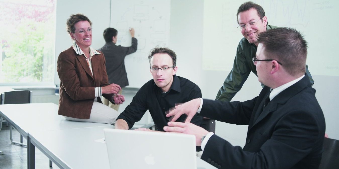 vier Personen besprechen sich, im Hintergrund eine Person am Whiteboard