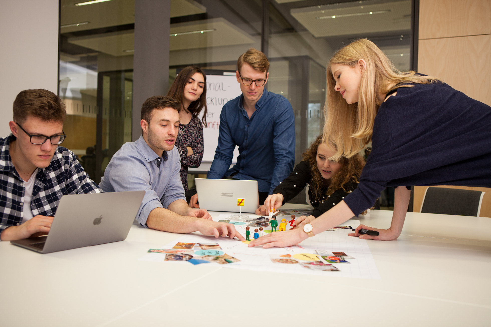 Gruppe von Studierenden sitzt und steht an einem Tisch mit Laptop und Spielsteinen in einem modernen Innenraum.